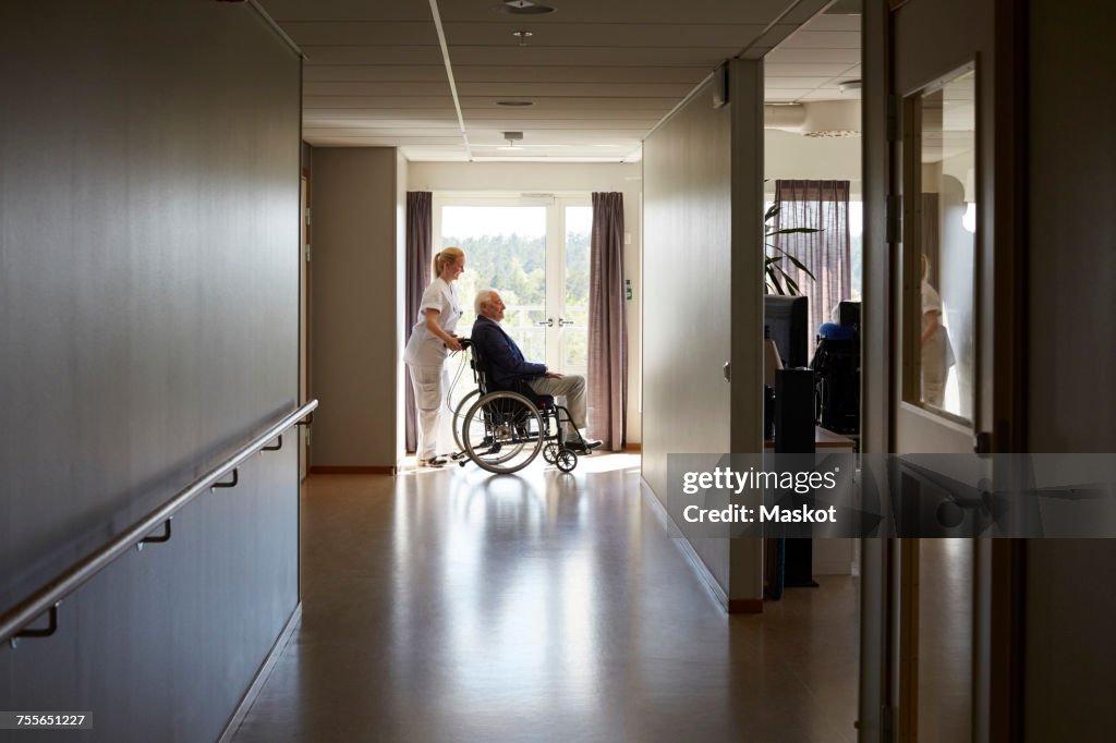 Full length side view of female nurse pushing senior man on wheelchair at hospital corridor