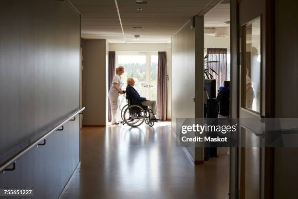 full length side view of female nurse pushing senior man on wheelchair at hospital corridor - senior care stock pictures, royalty-free photos & images