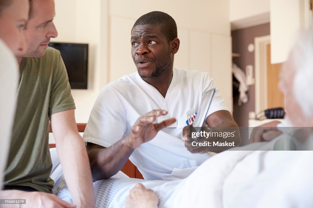 Male nurse discussing over digital tablet with couple while senior man reclining at hospital ward