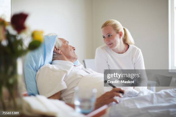 daughter talking to senior man reclining on hospital bed - family on bed stock-fotos und bilder