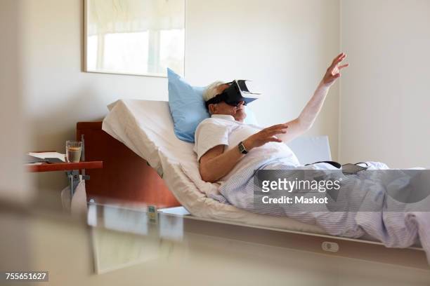 senior man gesturing while using vr glasses on bed in hospital ward - virtual healthcare stock pictures, royalty-free photos & images