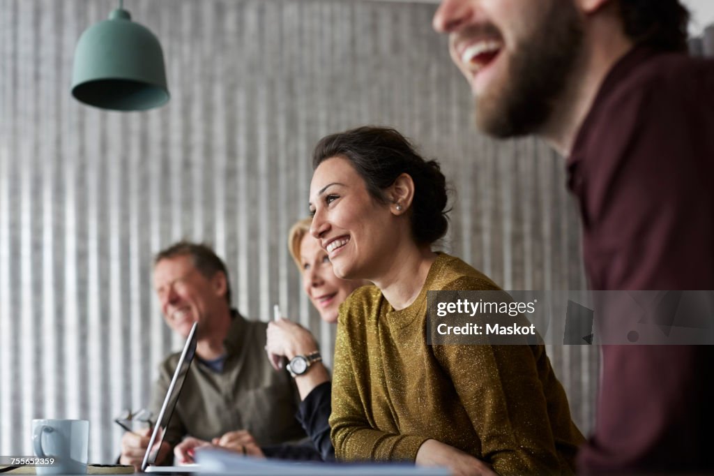 Cheerful creative business colleagues sitting at conference table while looking away in board room