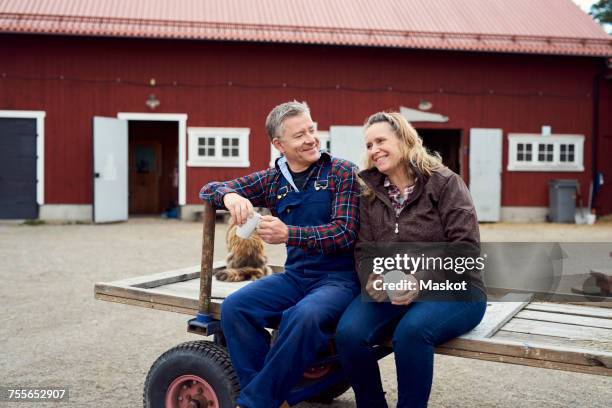 couple holding mugs while sitting on old-fashioned trailer against barn - couple farm photos et images de collection