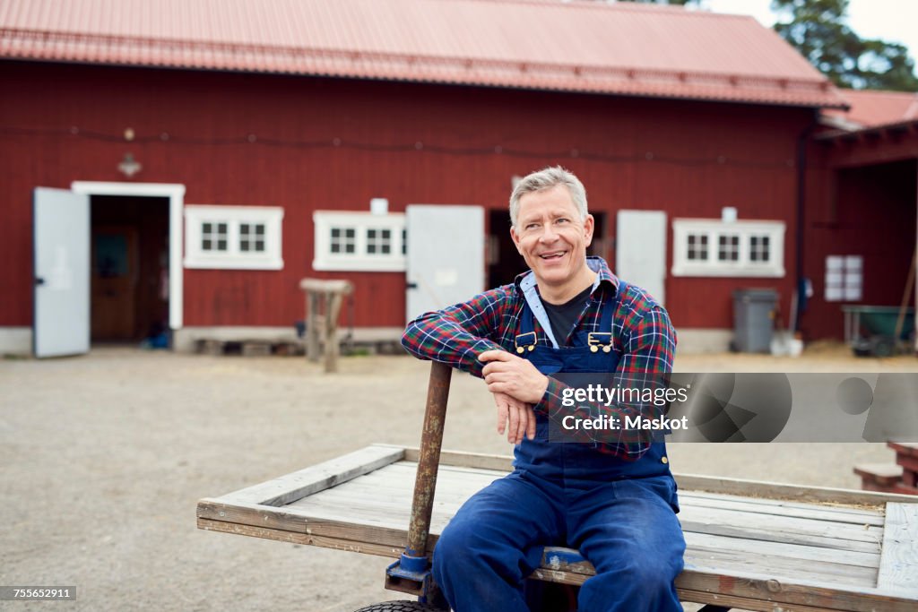 Happy farmer looking away while sitting on trailer against barn