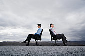 Two men sitting in office chairs outdoors with their backs against one another