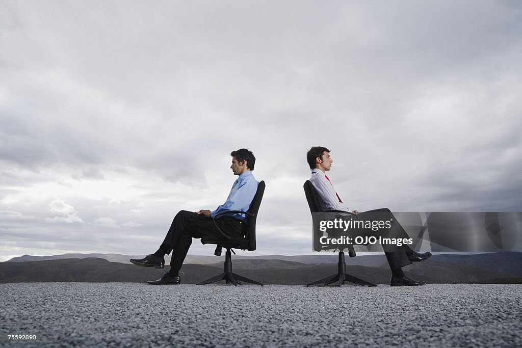 Two men sitting in office chairs outdoors with their backs against one another