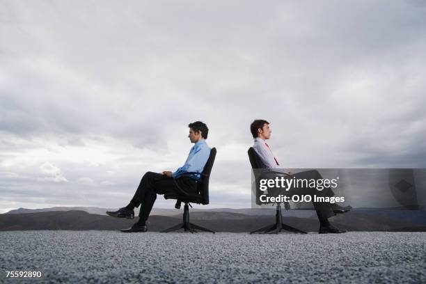 two men sitting in office chairs outdoors with their backs against one another - confrontation stock pictures, royalty-free photos & images