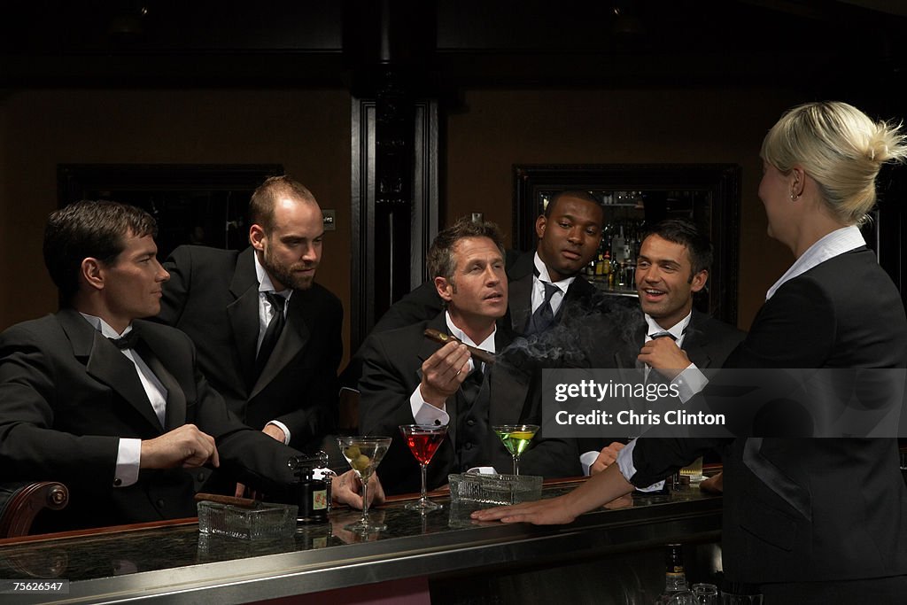 Men in dinner jackets drinking cocktails in bar, one holding cigar