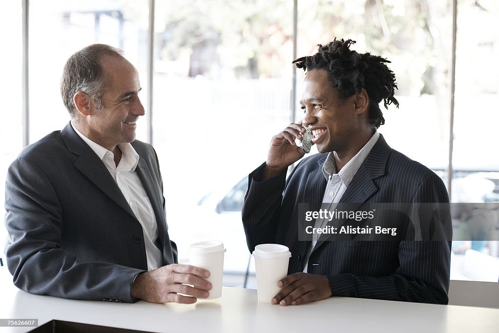 Two business men at bar counter holding coffee, smiling, one talking on mobile phone