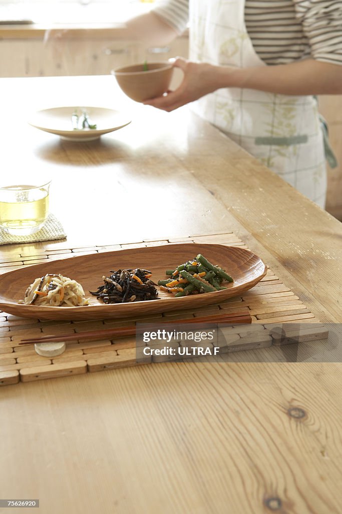 Japanese style vegetables in wooden tray with chopsticks on wooden table, woman preparing food in background