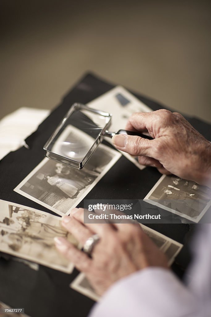 Senior woman looking at old photographs in album through magnifying glass, close-up of hands