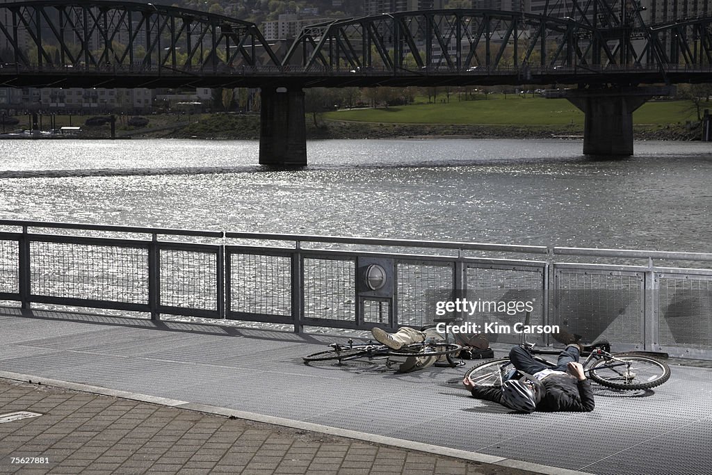 Two men with bicycles lying on pavement by river after collision