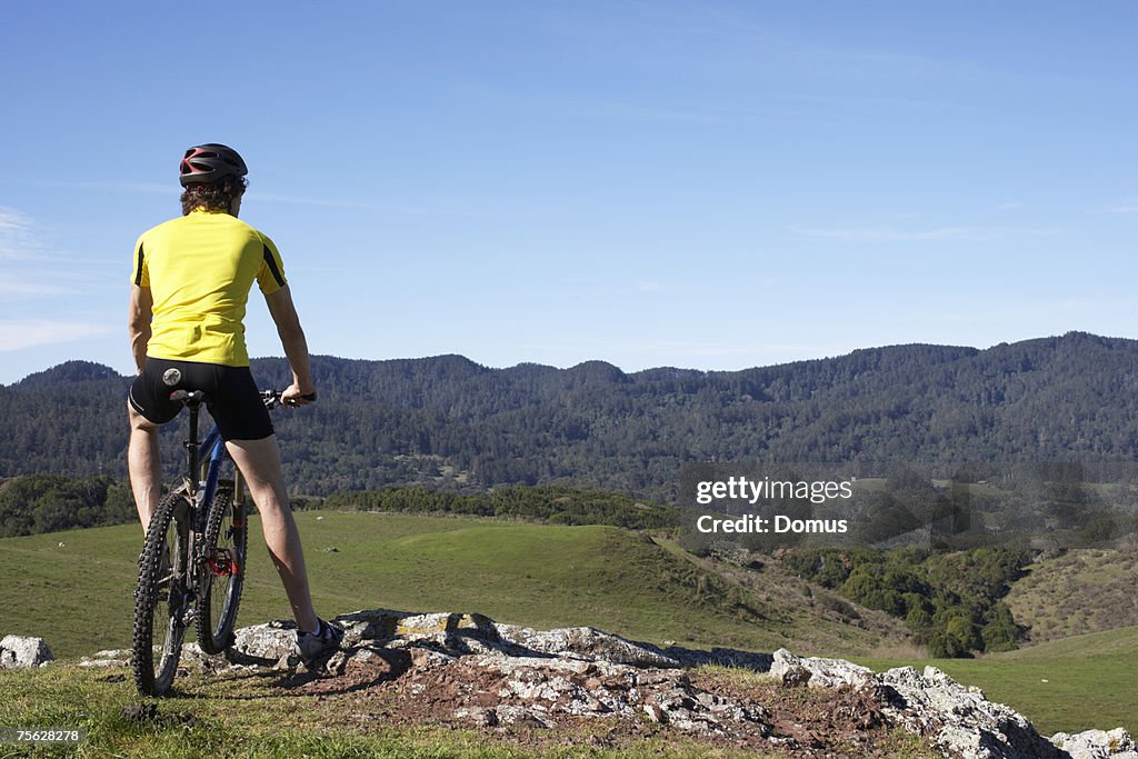 Man on mountain bike standing at edge of rock, looking at landscape, rear view