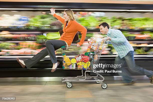 man pushing woman in supermarket trolley down aisle, blurred motion - shopping trolley photos et images de collection