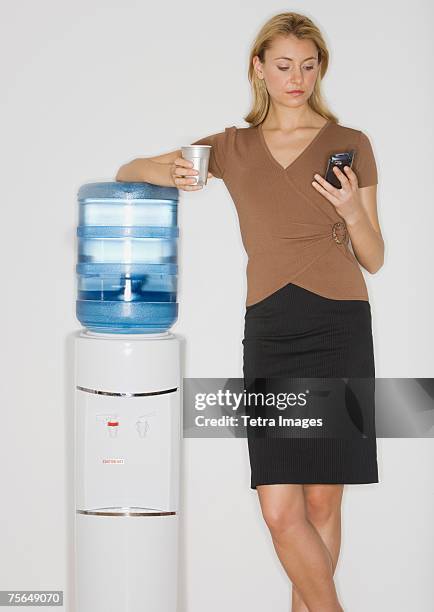 businesswoman looking at cell phone next to water cooler - water cooler white background stock pictures, royalty-free photos & images