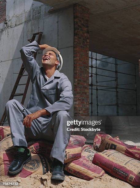 construction worker sitting on cement sack, looking up, laughing - sacchi cemento foto e immagini stock