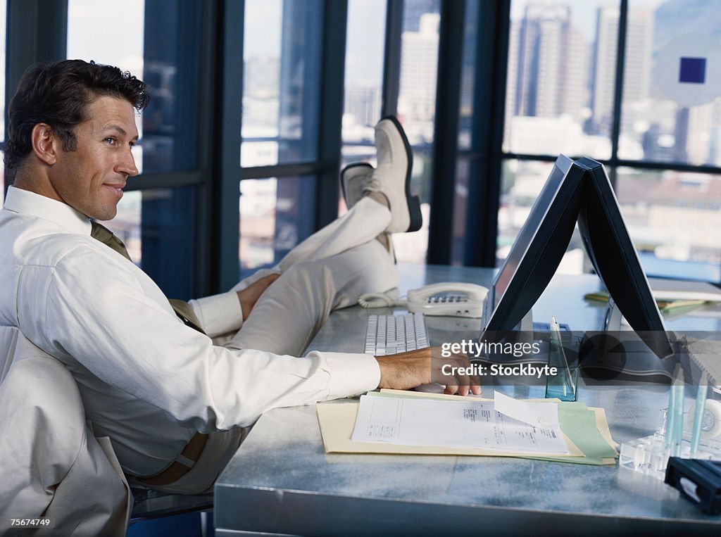 Businessman sitting with legs on table, using computer, smiling