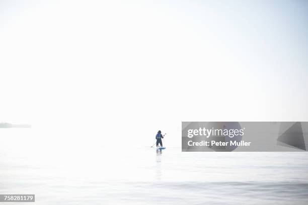 distant view of female paddleboarder paddling out on misty sea - high key stock pictures, royalty-free photos & images