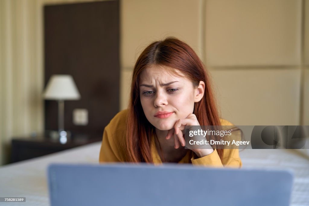 Confused Caucasian woman laying on bed using laptop