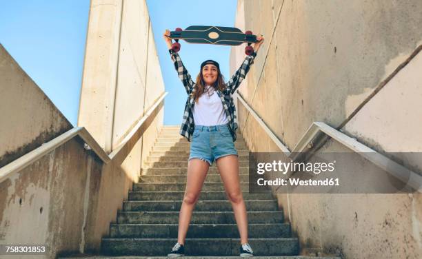portrait of happy young woman standing on staircase holding up her longboard - women in daisy dukes stockfoto's en -beelden