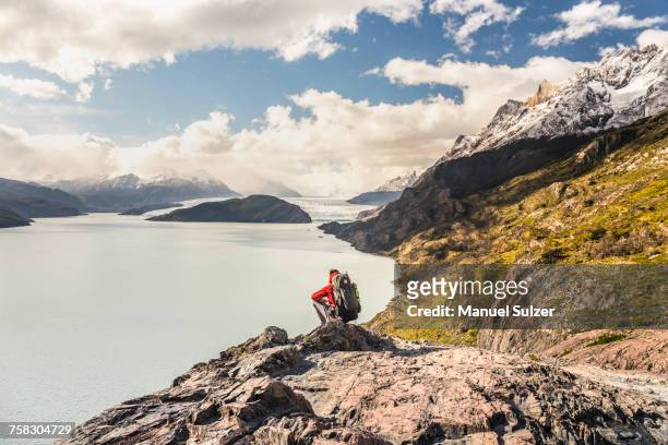 male hiker crouching to look out over grey lake and glacier, torres del paine national park, chile - puerto natales stock-fotos und bilder