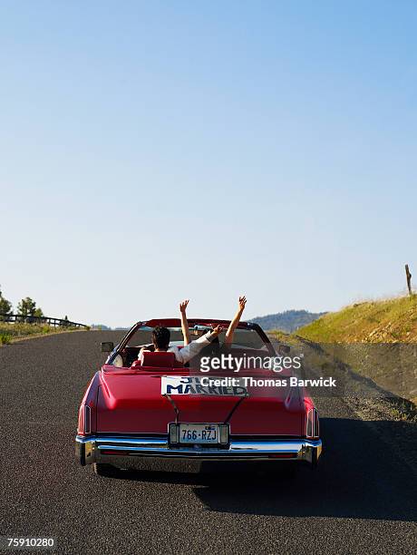 newlyeds driving down rural road in convertible with 'just married' sign, arms raised - just married car stockfoto's en -beelden