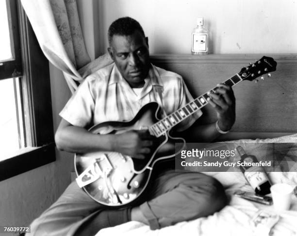 Blues musician Howlin' Wolf poses for a portrait in a hotel room holding a hollowbody electric guitar and sitting on a bed with a bottle of Ancient...