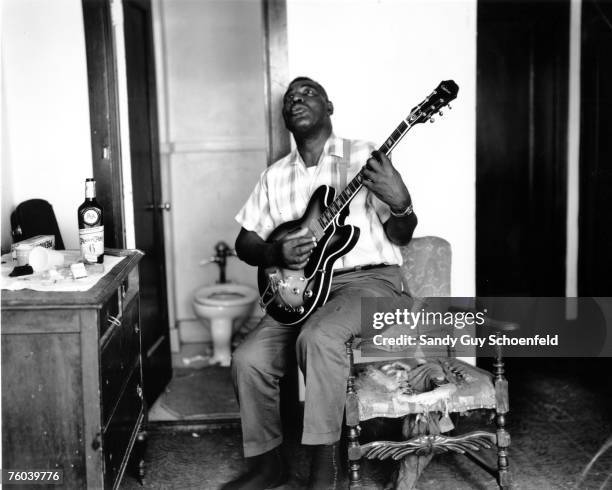 Blues musician Howlin' Wolf poses for a portrait in a hotel room holding a hollowbody electric guitar and sitting on a bed with a bottle of Ancient...