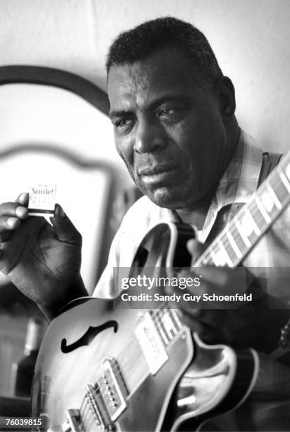 Blues musician Howlin' Wolf poses for a portrait holding a box of matches that reads "Now Smile" and a hollowbody electric guitar in a hotel room in...