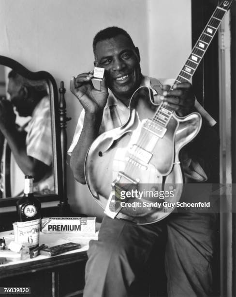 Blues musician Howlin' Wolf poses for a portrait holding a box of matches that reads "Now Smile", next to a bottle of Ancient Age bourbon, a box of...