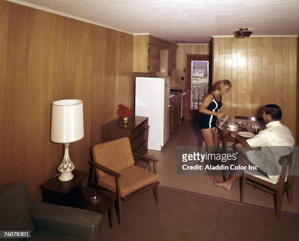 Smiling woman in a one-piece bathing suit pours a drink for a smiling man who sits at the table in the kitchenette of a wood-panelled room at an...