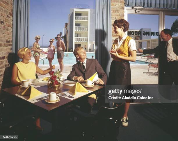 While several people in swimwear watch from outside the floor-to-ceiling windows, a smiling couple place an order with a smiling, uniformed waitress...