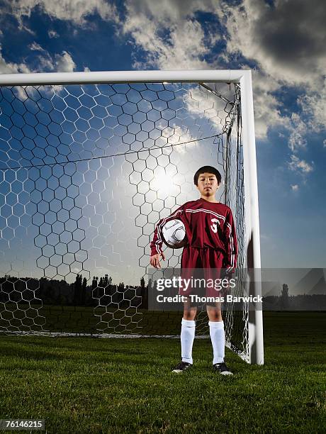 boy (10-11) playing goaltender, standing in goal holding football - boy goalie standing in front of goal net stock pictures, royalty-free photos & images