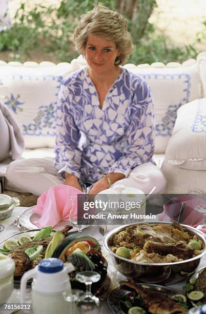 Diana, Princess of Wales, wearing a blue and white silk tunic with white silk trousers designed by Catherine Walker, sits cross-legged on cushions...