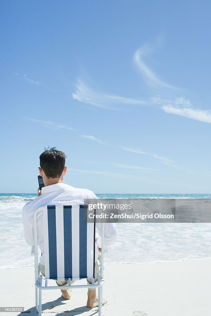 Businessman sitting in folding chair, holding up cell phone, on beach