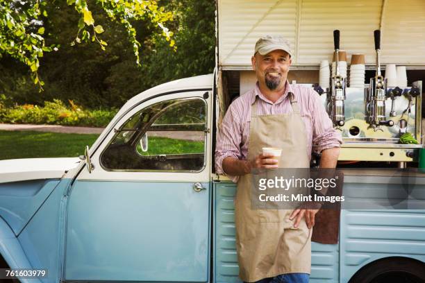 Bearded man wearing apron standing by blue mobile coffee shop, holding hot drink, smiling at camera.