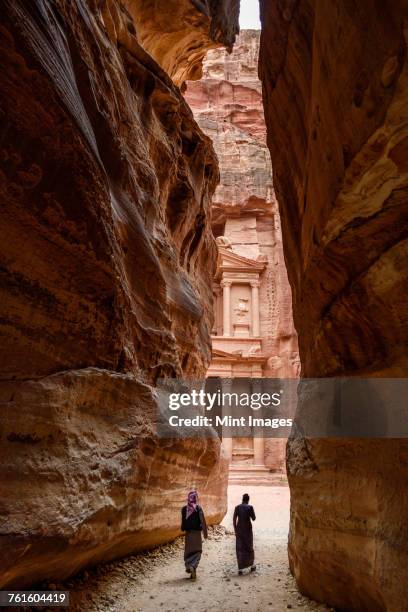 two people walking through narrow rock formation towards rock-cut architecture of al khazneh or the treasury at petra, jordan. - jordan country stock pictures, royalty-free photos & images