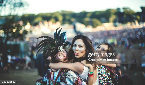 three young women at a summer music festival feather headdress and faces painted, smiling at camera, sticking out tongue. - body art stockfoto's en -beelden