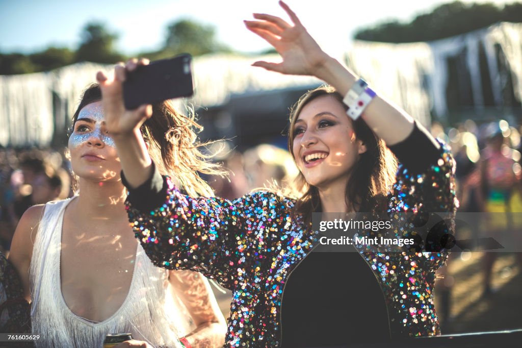 Young smiling woman at a summer music festival wearing multi-coloured sequinned jacket, taking picture with smartphone.