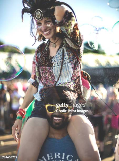 revellers at a summer music festival young man wearing yellow sunglasses carrying woman wearing feather headdress on his shoulders. - music festival 2016 weekend 2 stock pictures, royalty-free photos & images