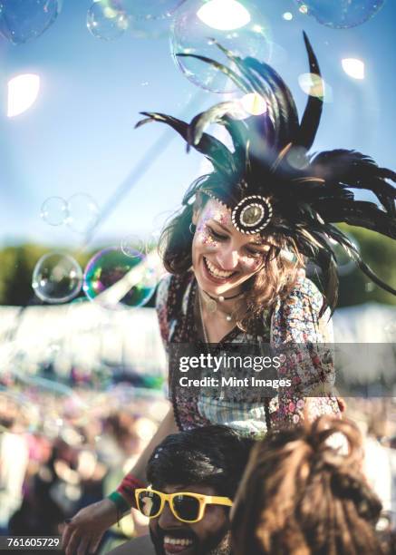 revellers at a summer music festival young man wearing yellow sunglasses carrying woman wearing feather headdress on his shoulders. - music festival 2016 weekend 2 photos et images de collection