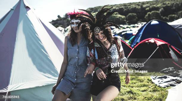 two young women at a summer music festival faces painted, wearing feather headdresses, walking arm in arm in between tents. - music festival 2016 weekend 2 stock pictures, royalty-free photos & images