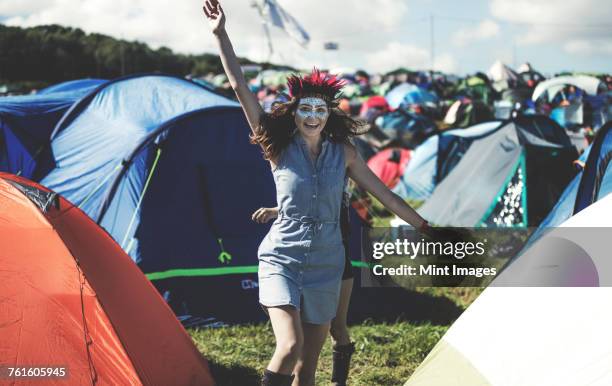 young woman at a summer music festival face painted, wearing feather headdress, standing near the campsite surrounded by tents, arm raised, smiling. - music festival day 1 stock-fotos und bilder