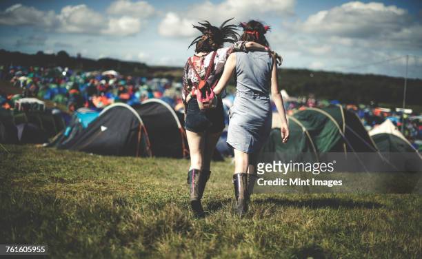 Rear view of two young women at a summer music festival wearing feather headdresses, walking arm in arm towards tents.