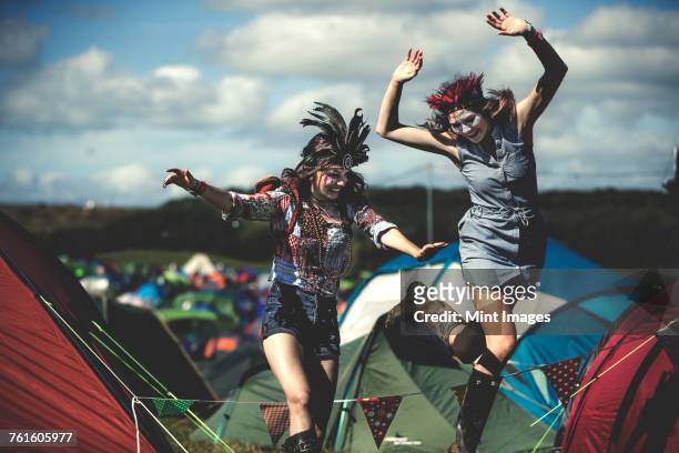 two smiling young women at a summer music festival face painted, wearing feather headdress, jumping among tents. - body painting femme bien etre photos et images de collection