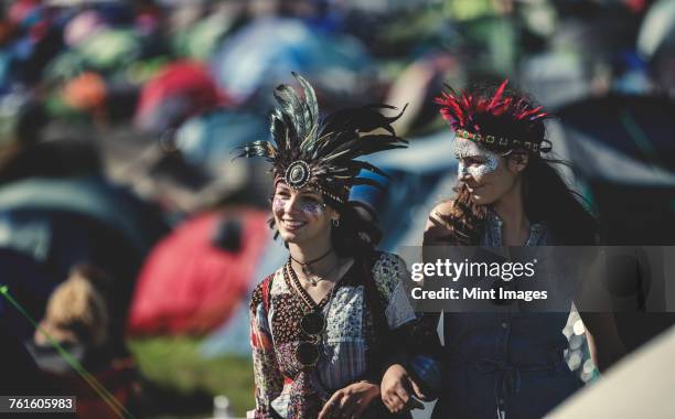 two smiling young women at a summer music festival face painted, wearing feather headdress, standing among tents. - music festival 2016 weekend 2 photos et images de collection