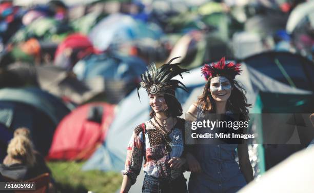 two smiling young women at a summer music festival face painted, wearing feather headdress, standing among tents. - music festival 2016 weekend 2 stock-fotos und bilder