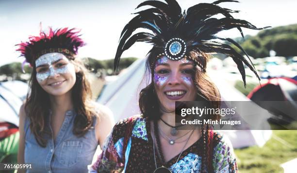 two smiling young women at a summer music festival face painted, wearing feather headdress, standing among tents. - music festival 2016 weekend 2 stock pictures, royalty-free photos & images