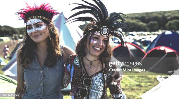 two smiling young women at a summer music festival face painted, wearing feather headdress, standing among tents. - music festival 2016 weekend 2 stock-fotos und bilder