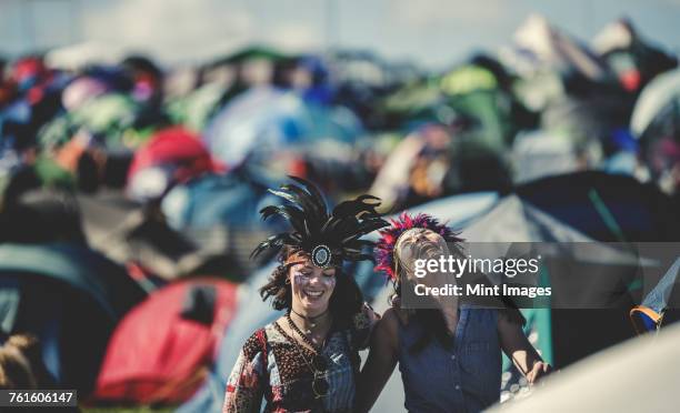 two smiling young women at a summer music festival face painted, wearing feather headdress, standing among the pitched tents. - music festival 2016 weekend 2 stock-fotos und bilder